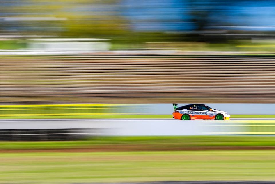 Orange and white race hatchback panned sharp against a motion-blurred grandstand