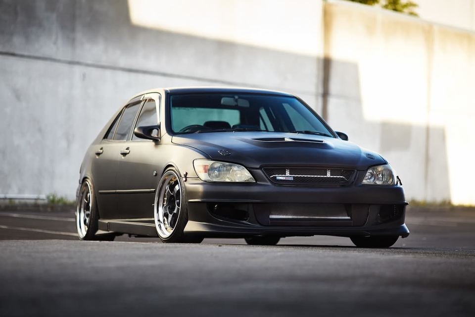 Dark grey sedan on deep-dish wheels parked against a concrete wall