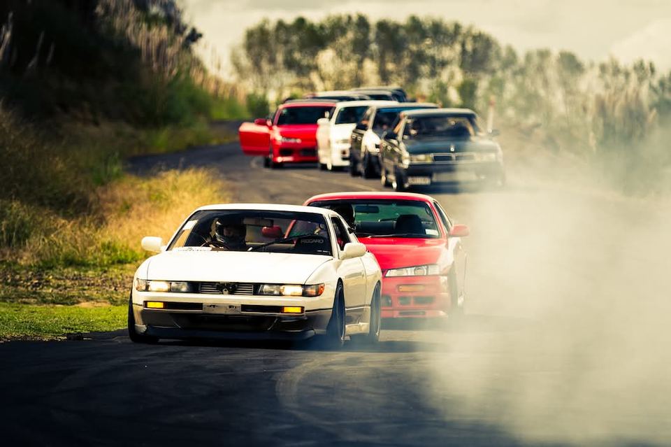 A line of drift cars nose to tail through dust, a white coupe leading