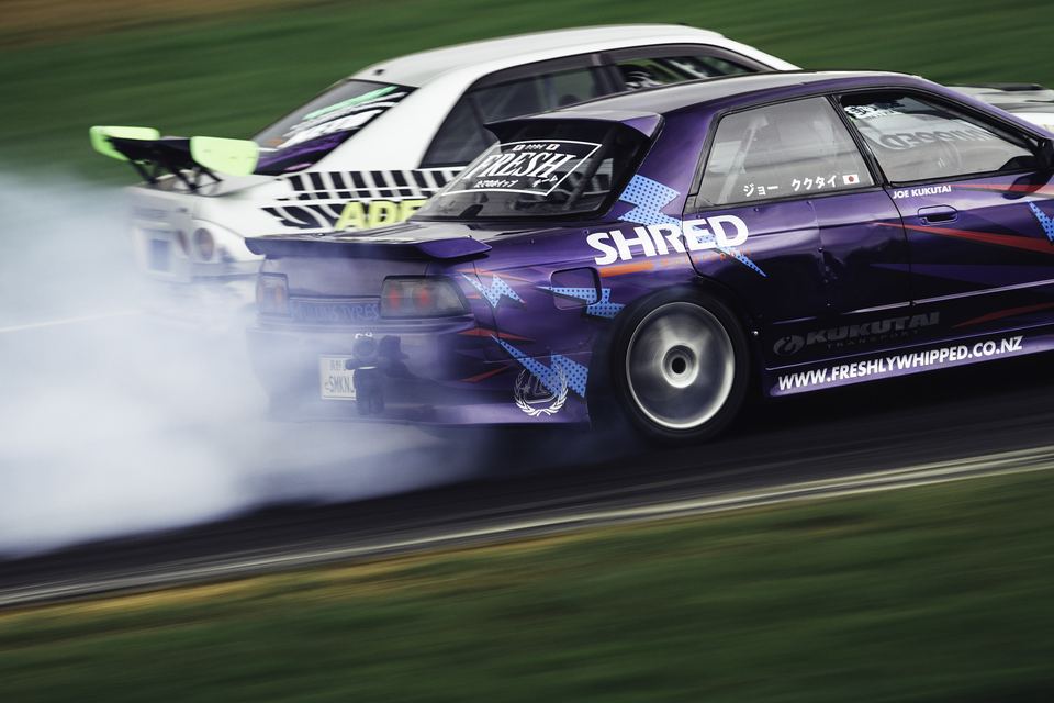 Purple and white drift car mid-slide trailing smoke across the grass