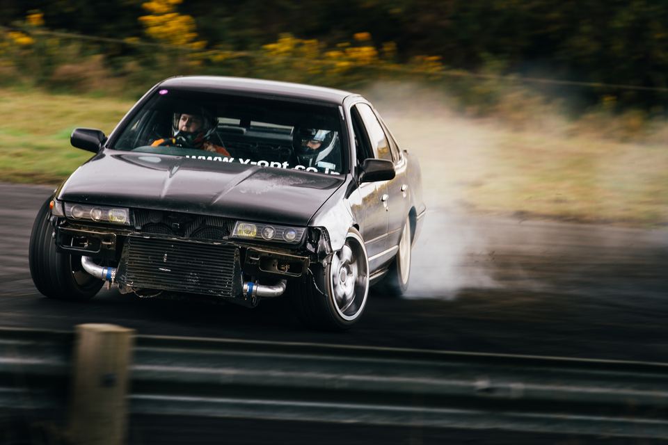 Silver sedan drifting toward the camera, front bumper missing, smoke behind