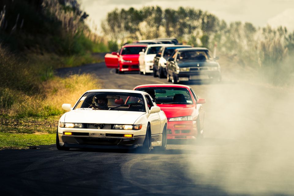 White coupe leading a train of drift cars through dust and smoke