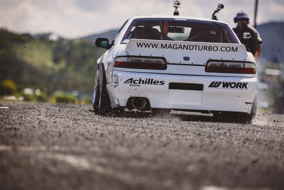White drift car with sponsor stickers seen from behind, kicking up gravel on the exit